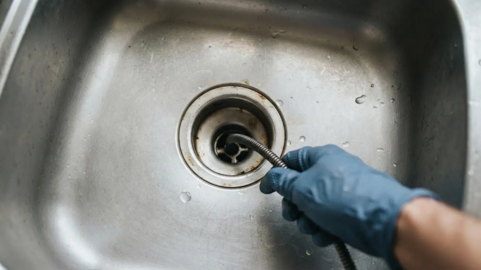 Gloved hand feeding a drain cable into a stainless steel kitchen sink drain